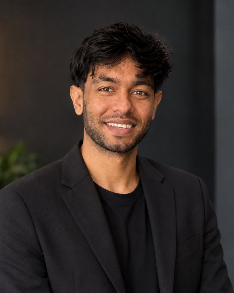 Abrar Khan, smiling, in a black blazer against a dark charcoal wall